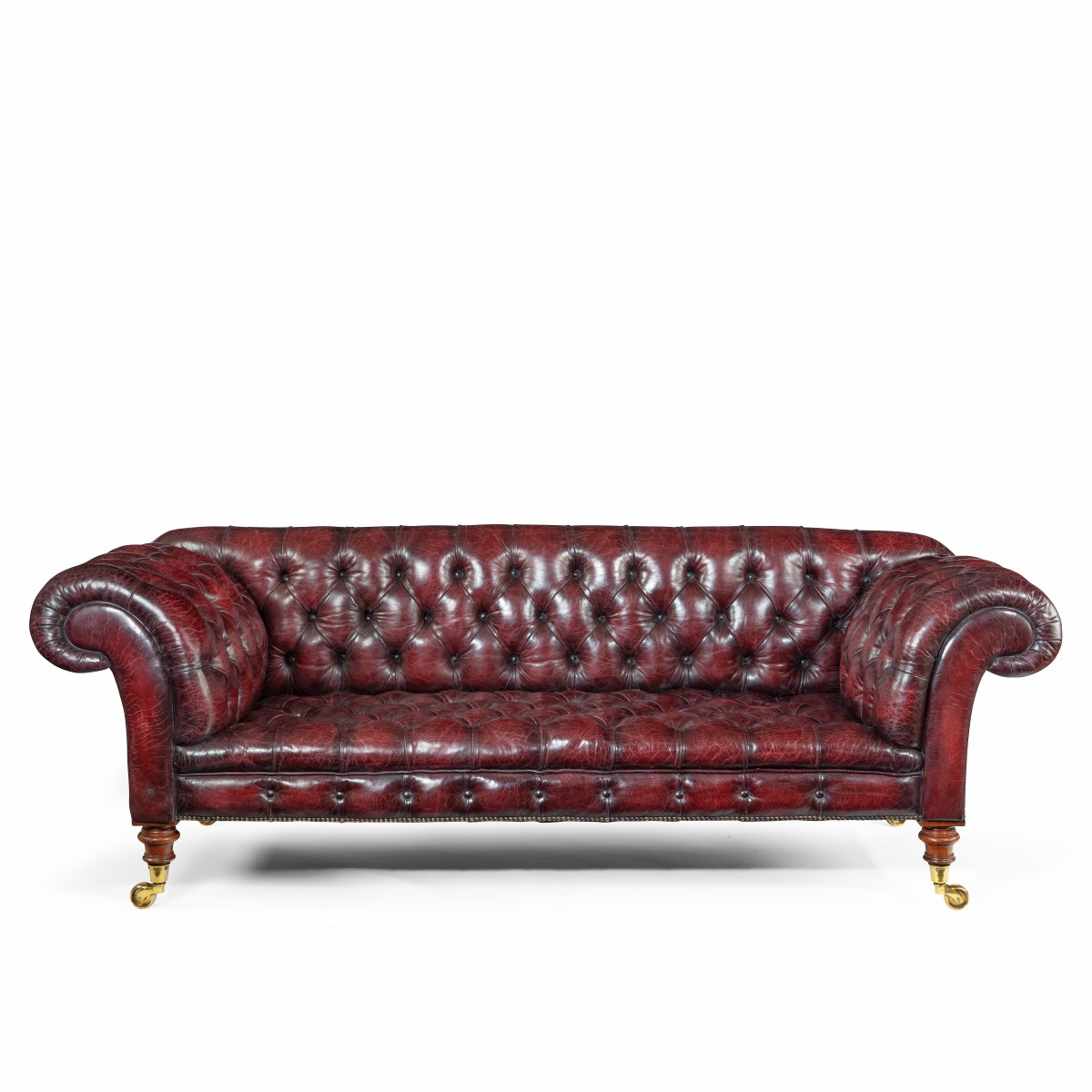 A Victorian deep buttoned Chesterfield settee, with an upright back and scroll arms, set on four turned walnut legs with brass castors, reupholstered in distressed burgundy leather