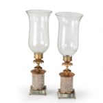 A pair of decorative storm lamps, each with a candle holder enclosed in a clear glass shade set on a turned gilt-brass support with a collar of petals and a cylindrical marble column, the rectangular base of striped marble. English, early 19th century.