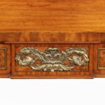 The breakfront rectangular top is raised upon four front and two back scroll legs carved with acanthus leaves and raised on hairy-paw feet. The panelled frieze encloses a disguised mahogany-lined central drawer and is decorated with brass stringing and a central foliate mount flanked by an athemion above each leg. English details