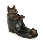 A Black Forest fruitwood carving of a cat in a boot, carved with a cat with inlaid glass eyes resting in the mouth of an ankle boot while the tips of his paws and tail peep out from the open toe of the boot, the boot stained black, the head hinged to form the lid.  Swiss, circa 1890.