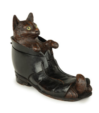 A Black Forest fruitwood carving of a cat in a boot, carved with a cat with inlaid glass eyes resting in the mouth of an ankle boot while the tips of his paws and tail peep out from the open toe of the boot, the boot stained black, the head hinged to form the lid.  Swiss, circa 1890.