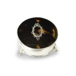 A small circular tortoiseshell piqué patch pot by Mappin and Webb, the hinged lid with a beribboned oval silver piqué escutcheon, raised on three fleur-de-lys feet. English, circa 1920, assay mark W, others indistinct.