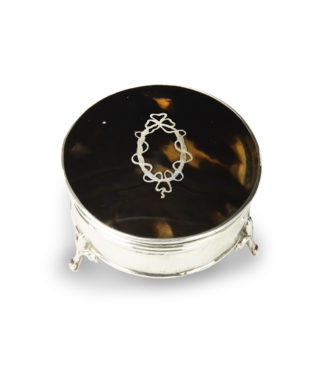 A small circular tortoiseshell piqué patch pot by Mappin and Webb, the hinged lid with a beribboned oval silver piqué escutcheon, raised on three fleur-de-lys feet. English, circa 1920, assay mark W, others indistinct.