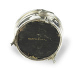 A small circular tortoiseshell piqué patch pot by Mappin and Webb, the hinged lid with a beribboned oval silver piqué escutcheon, raised on three fleur-de-lys feet. English, circa 1920, assay mark W, others indistinct Under