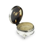 A small circular tortoiseshell piqué patch pot by Mappin and Webb, the hinged lid with a beribboned oval silver piqué escutcheon, raised on three fleur-de-lys feet. English, circa 1920, assay mark W, others indistinct hallmarked open