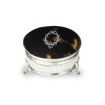 A small circular tortoiseshell piqué patch pot by Mappin and Webb, the hinged lid with a beribboned oval silver piqué escutcheon, raised on three fleur-de-lys feet. English, circa 1920, assay mark W, others indistinct hallmarked