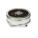 A small oval tortoiseshell piqué patch pot, the hinged lid with a beribboned garland of flowers round a blank escutcheon, raised on four ogee feet. English, assay marks C&A, London, 1919.