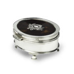 A small oval tortoiseshell piqué patch pot, the hinged lid with a beribboned garland of flowers round a blank escutcheon, raised on four ogee feet. English, assay marks C&A, London, 1919 -
