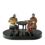 A rare Anglo-Chinese Regency polychrome painted wax and wood group of two Chinese card players, both with nodding heads with horsetail top-knots, wearing jackets with large collars, one with wide-sleeves,  gilt belts and voluminous trousers, seated at a circular table, one man grinning and pointing in amusement at the frustration of his companion at having dropped his cards on the floor.  Anglo-Chinese