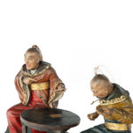 A rare Anglo-Chinese Regency polychrome painted wax and wood group of two Chinese card players, both with nodding heads with horsetail top-knots, wearing jackets with large collars, one with wide-sleeves,  gilt belts and voluminous trousers, seated at a circular table, one man grinning and pointing in amusement at the frustration of his companion at having dropped his cards on the floor